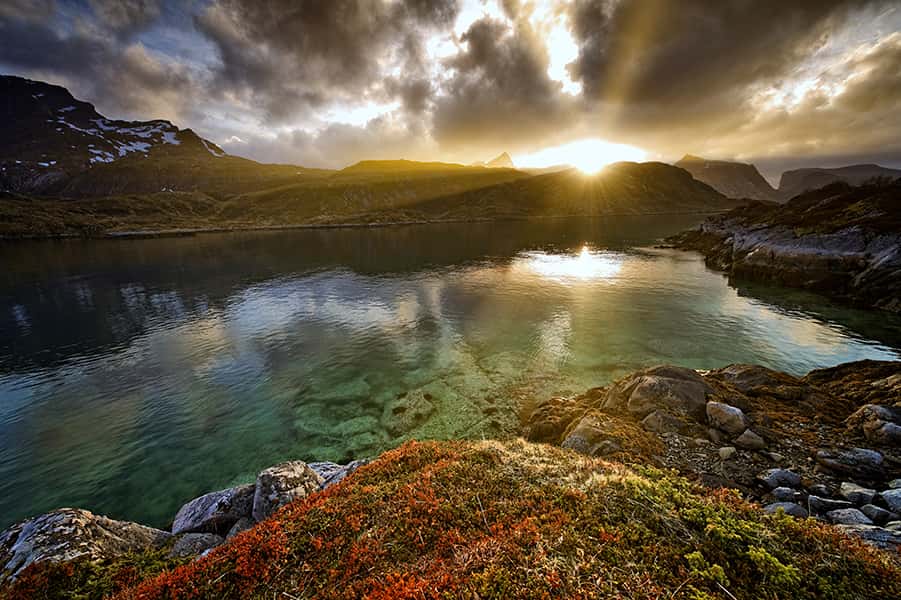 Die Sonne erstrahlt hinter den Bergen auf einen See auf den Lofoten