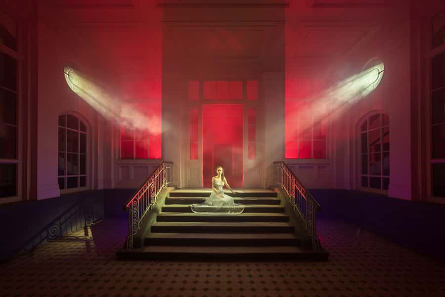 zwei Lichter scheinen auf eine Frau auf einer Treppenstufe, dahinter rotes Licht, in einem Raummilieu
